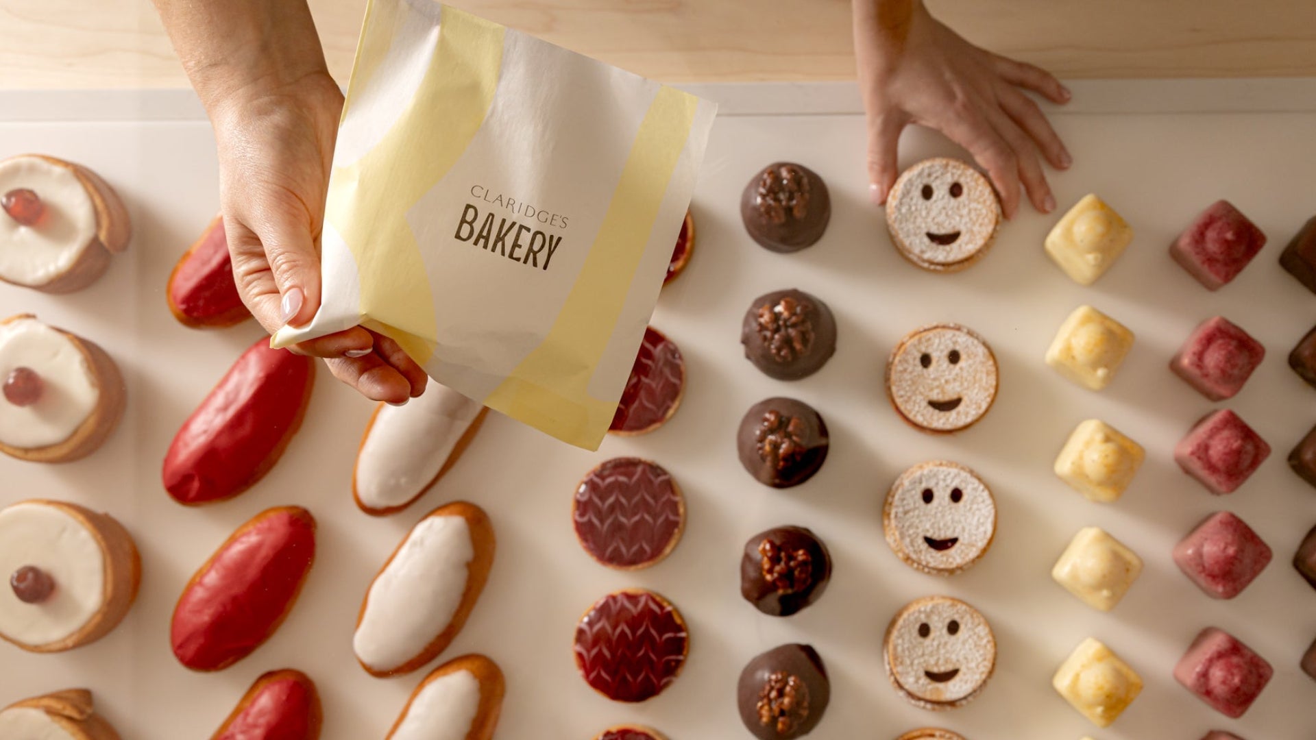 Hands holding a bag from Claridge's Bakery with various small baked goods on a light surface.