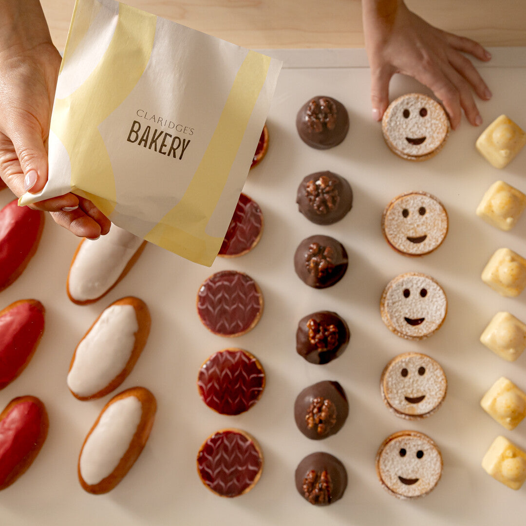 Hands holding a bag from Claridge's Bakery with various small baked goods on a light surface.