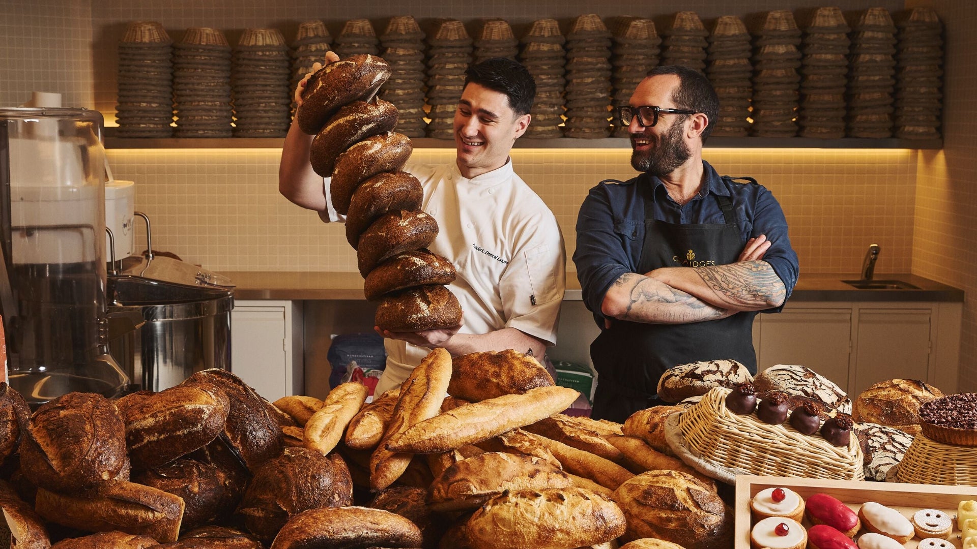 Two bakers standing behind a counter with various bread loaves in a bakery.