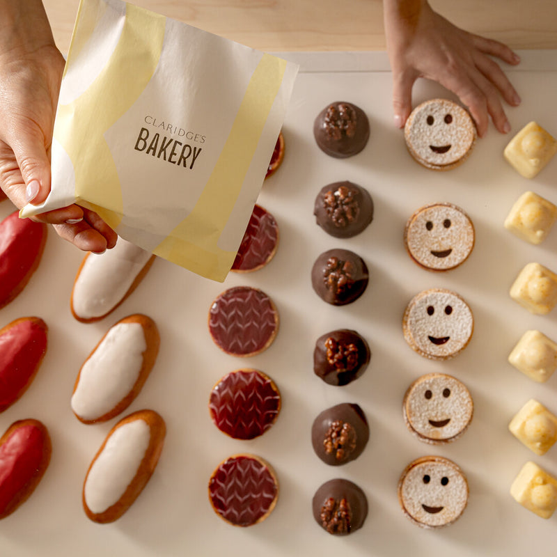 Hands holding a bag from Claridge's Bakery with various small baked goods on a light surface.